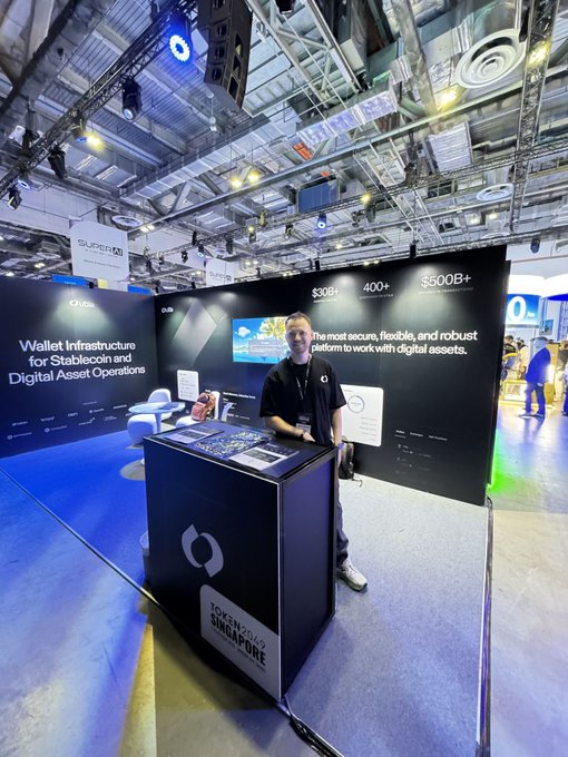 A man wearing a mask stands behind an "IOX125 X SINGAPORE" display booth at a conference, promoting "Wallet Infrastructure for Stablecoin and Digital Asset Operations" with statistics showing $30B+ and 400+.