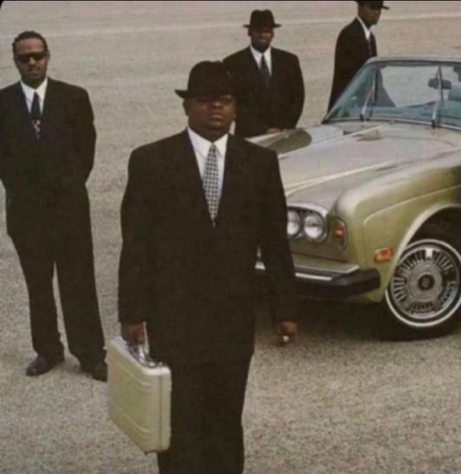 Three men in dark suits and hats stand near a luxury car, with the central figure carrying a silver briefcase, evoking a bodyguard or gangster aesthetic.