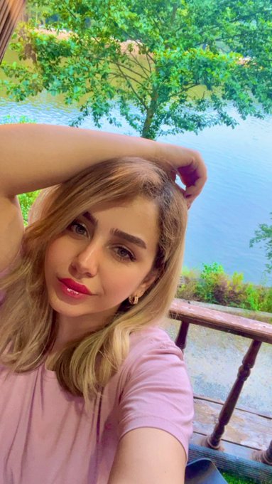A woman with blonde hair in a pink t-shirt poses with her hand on her head near a window overlooking water and trees.