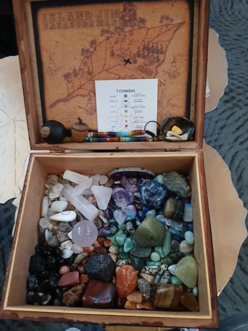 An open wooden treasure box from 'ISLAND JIM'S TREASURE MAP' containing an assortment of colorful tumbled stones and crystals, with a card showing the 7 chakras positioned against a vintage-style map on the inner lid.