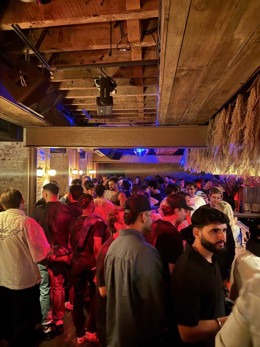 A crowded, dimly lit venue with wooden ceiling beams and red and blue lighting, where many people are gathered close together.
