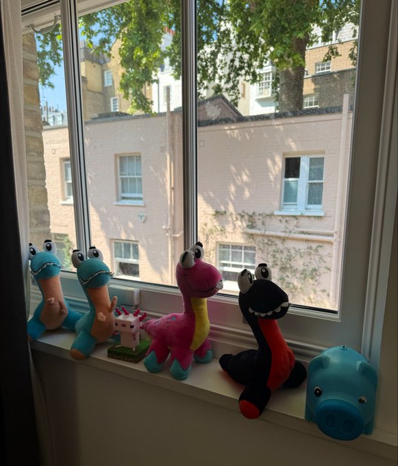 A windowsill display of colorful plush dinosaur toys and a small house figurine, looking out onto brick buildings and trees.