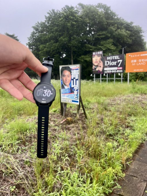 A hand holds a Garmin smartwatch displaying "01:30" against a backdrop of overgrown grass and political/campaign billboards, including one reading "HOTEL Dior 7." The image humorously illustrates a lost Garmin watch discovered in the field, connecting to the post about losing the device shortly after changing the strap in the #STEPN fitness app.