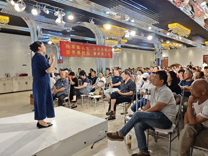 A woman in a blue dress speaks to a seated audience at what appears to be a promotional event, with a Chinese banner reading "结苹果股东 享苹果人生" (Become an Apple shareholder, enjoy the Apple life) visible behind her. The image illustrates the speaker's point about how such grassroots marketing schemes, which traditionally promoted scam crypto tokens, could pivot toward promoting legitimate stock investments like Apple's shares.