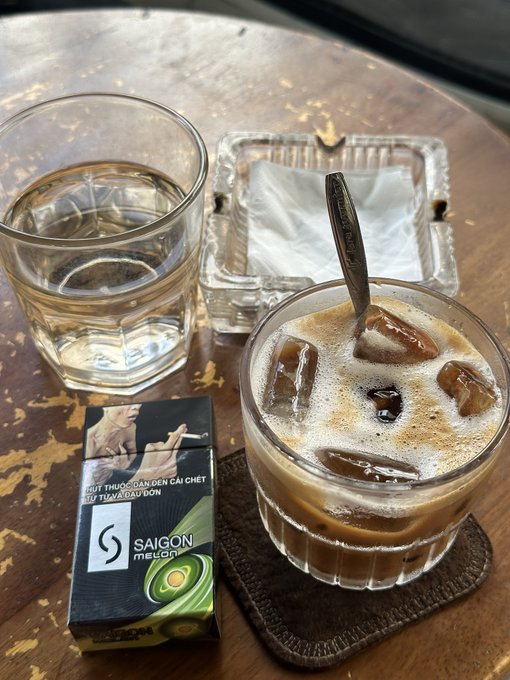 A glass of iced coffee and a glass of water sit on a wooden table alongside a pack of "Saigon" cigarettes with the Vietnamese text "HÚT THUỐC LÀ BẠN ĐẾN CÁI CHẾT" (Smoking leads to death). The relaxed café scene pairs perfectly with the post's playful complaint about feeling lazy on the weekend—much like Bitcoin's sluggish movement, the vibe is unhurried and chill.