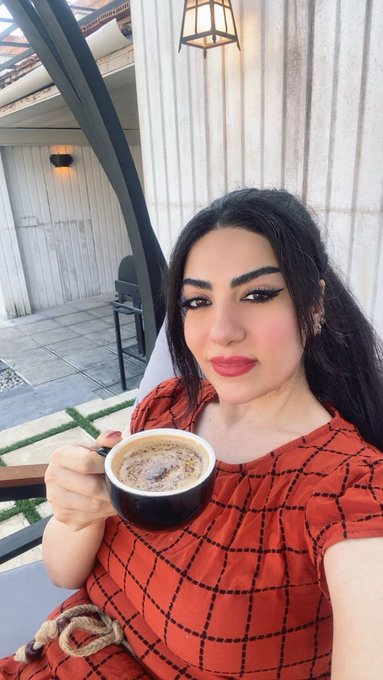 A woman with dark hair and bold eyeliner holds a cup of coffee while sitting in a sunlit patio with white wooden walls.