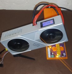 A photograph showing a silver electronic device with two speakers and an LCD screen connected to a brightly lit yellow circuit board, resting on a brown surface.