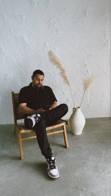 A man sits casually in a wooden chair beside a vase of pampas grass, dressed in dark clothing and sneakers while looking down at his phone. The image pairs with a post asserting South Asian, Pakistani pride and calling for an end to racism in crypto spaces—echoing the call for unity and humanity over prejudice.