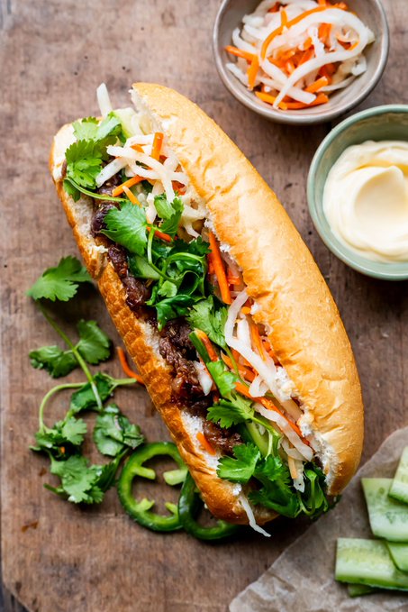 An open banh mi-style sandwich filled with meat, cilantro, pickled carrots and daikon, and jalapeños, accompanied by small bowls of extra pickles and mayonnaise on a wooden surface.