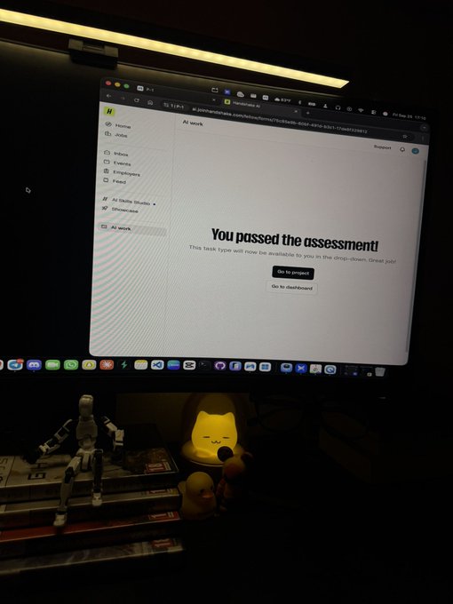 A computer monitor displays "You passed the assessment!" on a TaskType screen, with a lit cat-shaped lamp and figurines visible on the desk below.
