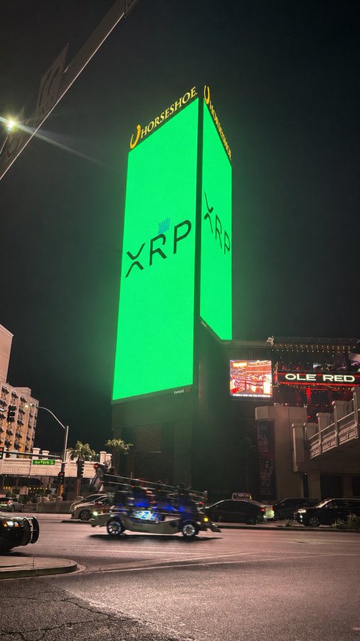 A towering illuminated billboard at the Horseshoe casino displays a bright green screen with "XRP" branding, while an "Ole Red" sign glows nearby. The image ties into the post's theme by symbolically showcasing XRP in a highly visible, celebratory manner, as if the entire structure is shouting the ticker to the Las Vegas night.