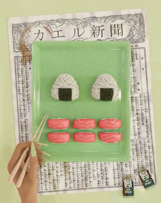 A green square plate holds two onigiri and six salmon sushi pieces arranged like a face, placed over a Japanese newspaper reading 'カエル新聞' (Frog Newspaper), with a hand holding chopsticks and two soy sauce packets labeled 'SOY SAUCE' beside it.