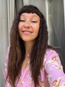 A woman with long dark hair and bangs smiling with her eyes closed, wearing a pink floral blouse, posing against a gray door.