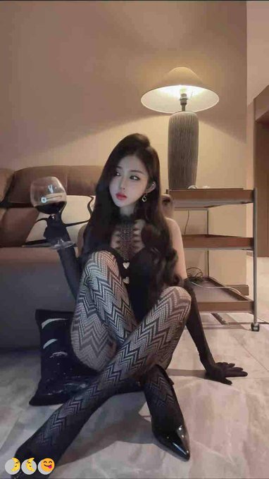 A young woman posing indoors on a light wood floor, wearing black lace tights and a black outfit, holding a glass of red wine, with a lamp and couch in the background.