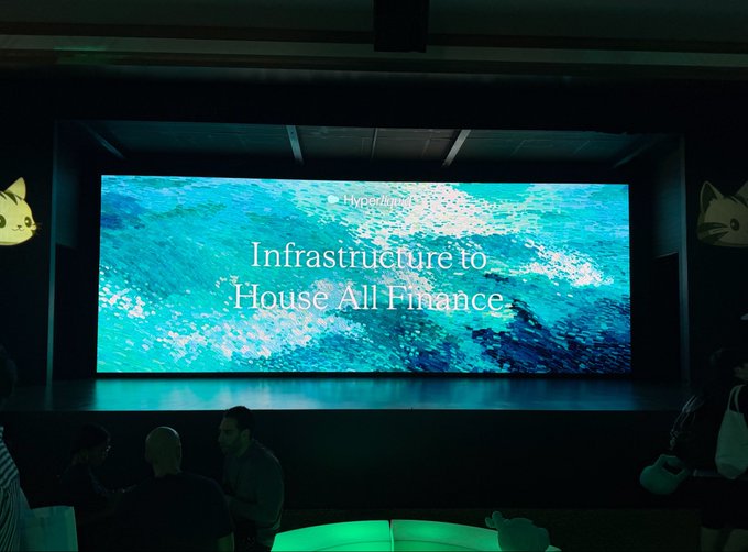 A presentation screen at a Hyperliquid event displays the text 'Infrastructure to House All Finance' over a blue water texture, with silhouettes of attendees and dog mascots visible below.