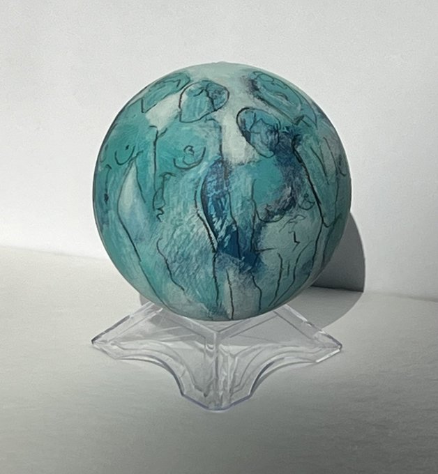 A round, light blue and green marbled object with abstract figure designs sits on a clear plastic display stand against a white background.
