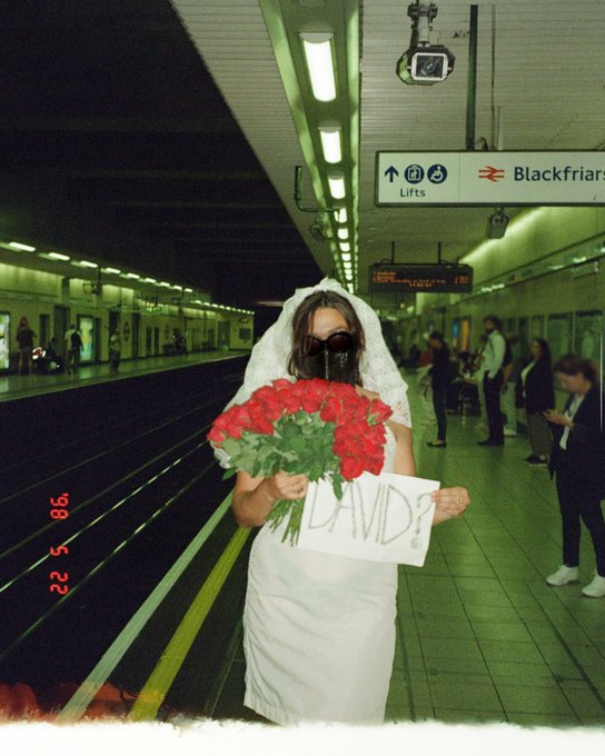 A woman wearing a white wedding dress, veil, and gas mask stands on a subway platform holding a bouquet of red flowers and a sign that says 'DAVID', with a sign for 'Blackfriar' visible behind her.