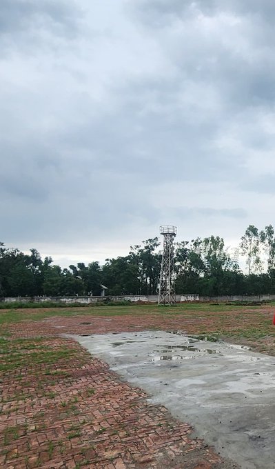 An outdoor scene under a cloudy gray sky showing a brick-paved area, a concrete path with puddles, trees, and a metal lattice tower in the distance.