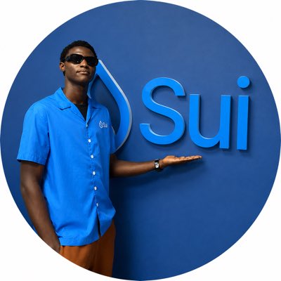 The image features a man wearing sunglasses standing next to a blue wall with the word "Sui" written on it. The man appears to be holding up his hand, possibly presenting something or engaging in conversation. The background of the photo is white, which contrasts with the blue wall and makes the text stand out more prominently.