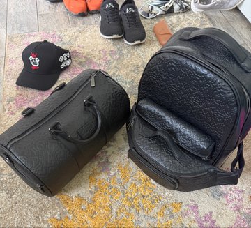 Two black quilted carry-on suitcases and a black baseball cap with logos sitting on a multi-colored patterned rug, with sneakers visible in the background.