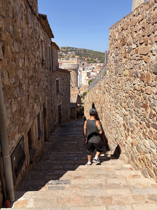 A person in a black tank top, shorts, and a cap walking up stone steps in a narrow alleyway between old stone buildings, with a view of a town and hills in the background.