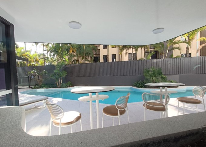 A modern covered outdoor entertaining area by a swimming pool features white round tables paired with wire-frame chairs and tan cushions, with apartment buildings and palm trees visible beyond a dark fence. The image showcases one of the poster's investment properties, illustrating the type of upscale residential amenity space that has appreciated and doubled in both rent and value since being purchased during the 2020 Covid downturn.