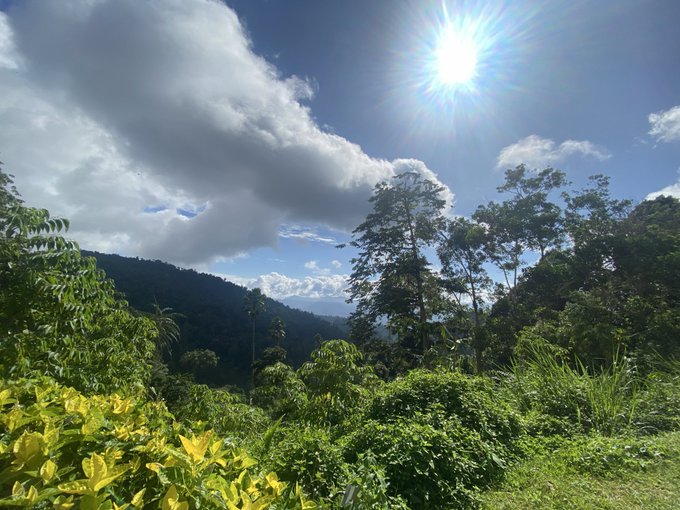 A scenic mountain landscape shows lush green vegetation under a bright sun and a blue sky filled with white clouds.