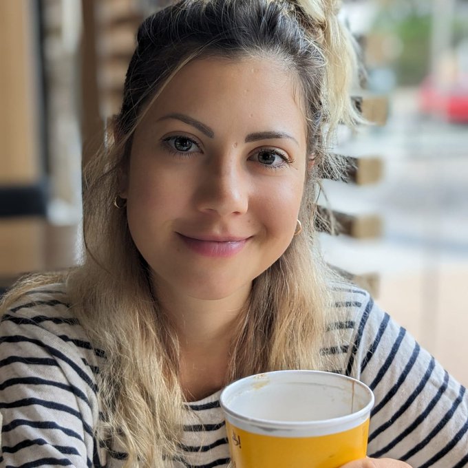 A blonde woman in a striped shirt holds a yellow coffee cup while taking a selfie, smiling softly at the camera. The casual, candid café setting reflects the post's message of unity and optimism in the crypto industry, suggesting a relaxed, approachable tone to the call for collaboration over division.