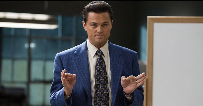 Leonardo DiCaprio in a blue pinstripe suit and patterned tie stands with his hands raised in a shrugging or explaining gesture, with a whiteboard visible to the right.