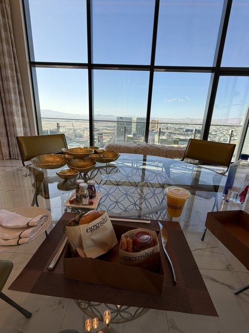 A meal service setup on a glass dining table includes Eggspert branded paper bags with food, a juice bottle, and condiment packets, all set in a high-rise room with panoramic city views through floor-to-ceiling windows.