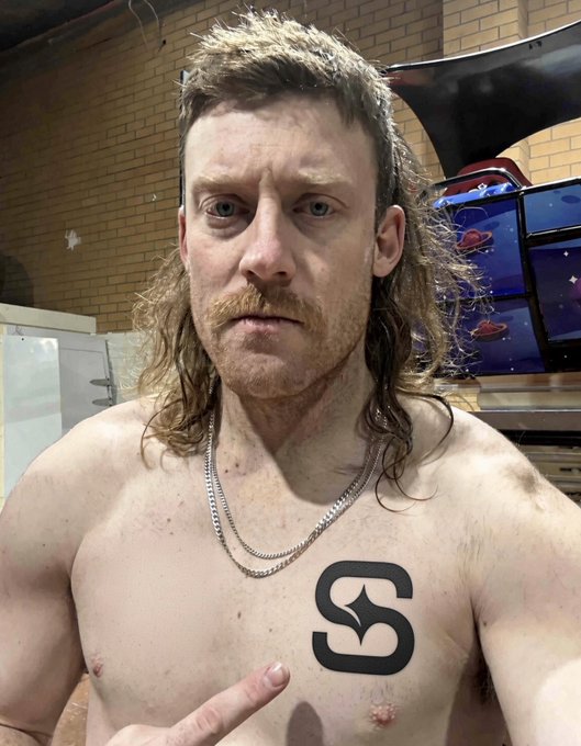 A shirtless man with a mullet and mustache taking a mirror selfie while pointing at an "S" logo sticker on his chest, with arcade machines visible in the background.