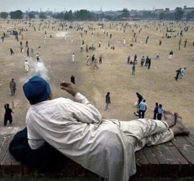 A man in white traditional clothing and a blue cap reclines on a brick ledge, smoking and looking out over a large dusty field filled with cricket players.