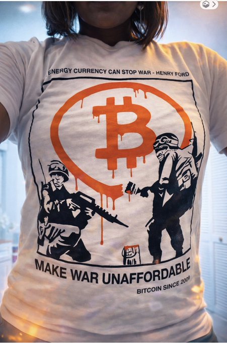 A person wears a white Bitcoin t-shirt reading "ENERGY CURRENCY CAN STOP WAR - HENRY FORD / MAKE WAR UNAFFORDABLE / BITCOIN SINCE 2009," featuring a dripping orange Bitcoin logo flanked by two armed figures, channeling the post's defiant "fuck the bears" celebration of stacking through the bear market with a ₿.