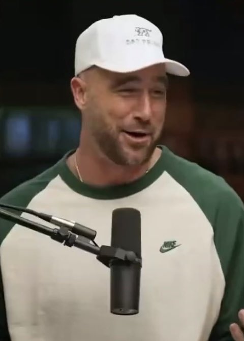 Travis Kelce, wearing a white cap and a green and white Nike sweatshirt, speaks into a microphone while smiling during an interview. The image accompanies his candid admission about getting away with a hold on Patrick Mahomes' rushing touchdown against the Broncos.
