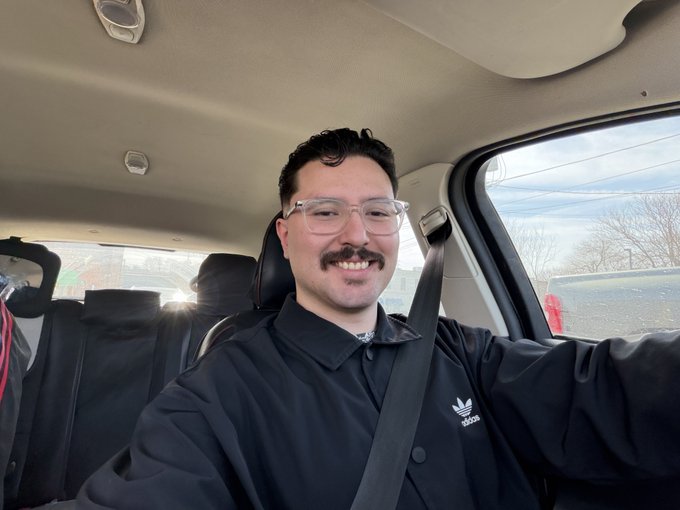Selfie of a smiling man with glasses and a mustache wearing a black Adidas jacket, seated in a car with a seatbelt on.