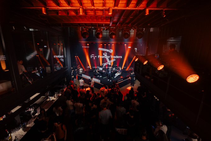 A crowd gathers in a dimly lit, modern event space bathed in red light, watching a presentation on a stage that prominently displays the "Frontier Traders" logo.