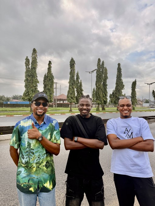 Three young men pose together on a road, smiling at the camera, with tall cypress trees and a cloudy sky in the background.