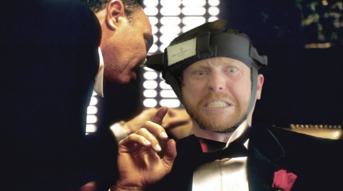 A man in a tuxedo grimaces while wearing a black head device, as another man in a suit leans in closely toward him.