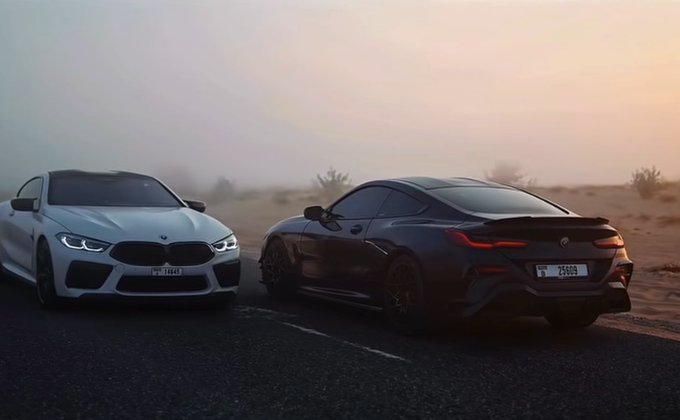 Two sleek BMW sports cars—one white and one black—park on a desert road at sunset, with license plates "T 74443" and "T 25899," evoking the post's humor about two friends celebrating together after striking it rich in crypto.