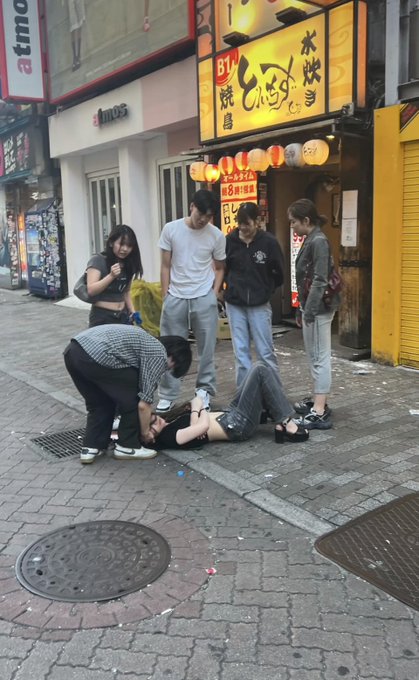 The image shows a group of people gathered around someone lying on the ground, with one person kneeling down next to them. They appear to be helping or attending to the individual who has fallen. The scene takes place on a city street, possibly near a store or restaurant, as indicated by the presence of a building and some signs in the background.