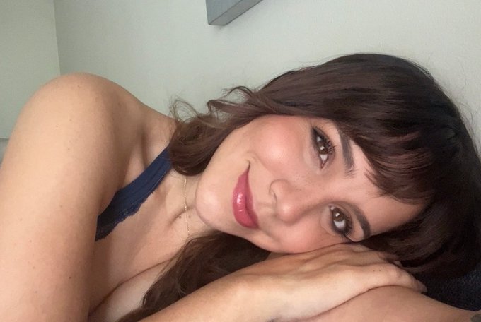 A woman with dark hair and bangs lies down resting her head on her arm, wearing a dark blue top, with glossy pink lips.