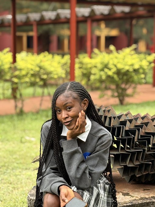 A young woman with long braided hair rests her chin on her hand while sitting outdoors, wearing a grey sweater and a plaid skirt next to a metal sculpture.