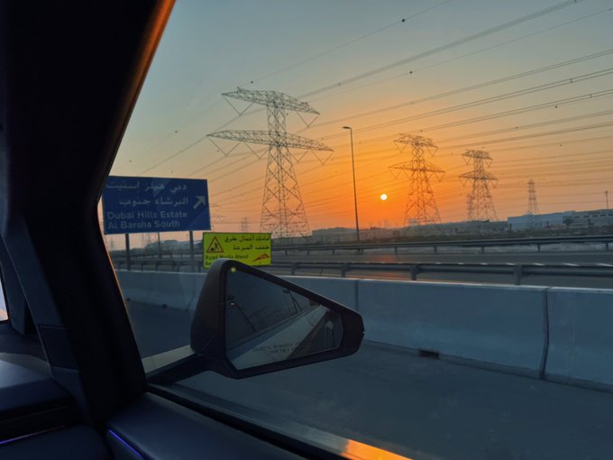 The image, taken from inside a car on a highway in Dubai, shows a sunset framed by electrical pylons and a road sign for "Dubai Hills Estate" and "Al Barsha South." It pairs with the post's message about choosing humility over online showboating—capturing a quiet, beautiful moment rather than a status symbol, since the poster has "nothing to flaunt."