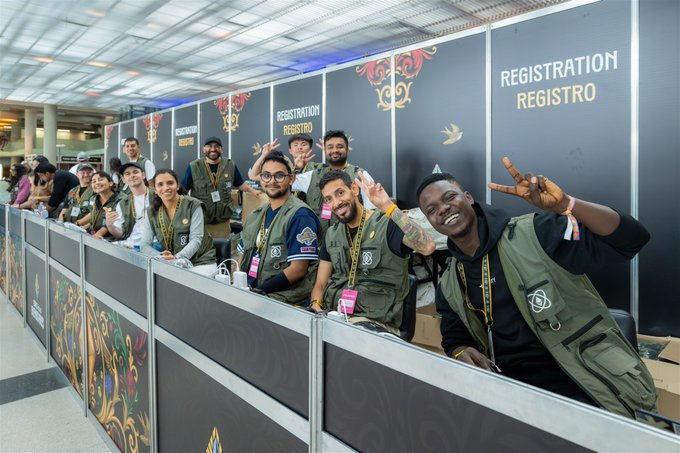 A group of smiling people in green volunteer vests posing behind a registration counter at an event, with 'REGISTRATION REGISTRO' visible on the dark backdrop.