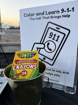 A photograph showing a box of Crayola Crayons (24 count) placed inside a gold-colored cup holder, with a printed paper in the background titled 'Color and Learn 9-1-1: The Call That Brings Help' featuring an illustration of a smartphone displaying 911.