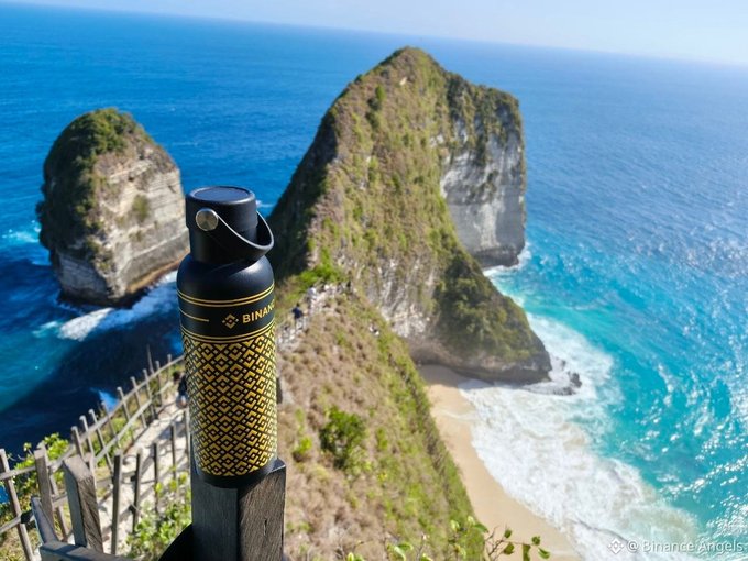 A Binance-branded black and yellow water bottle sits atop a wooden railing overlooking the dramatic cliff formations and turquoise waters of Kelingking Beach on Nusa Penida island, Indonesia, enjoying what the post claims is "the best view."