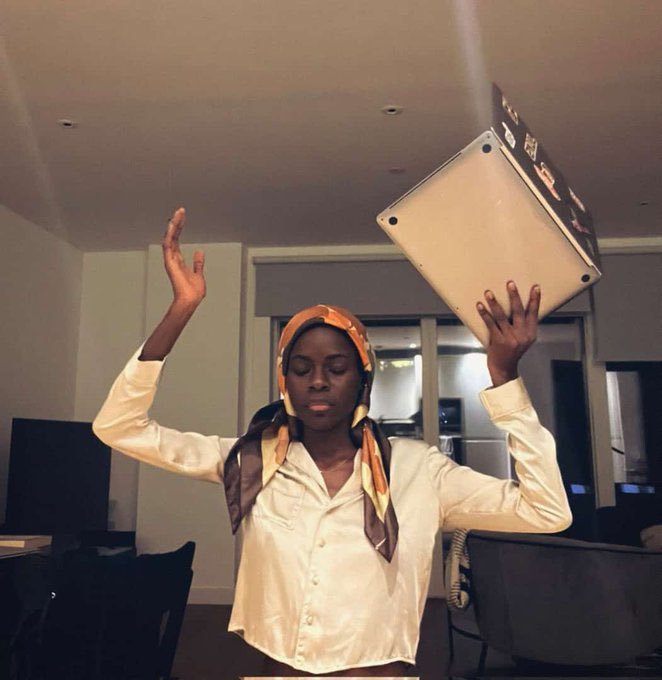 Photo of a young person wearing a white shirt and an orange patterned head wrap, posing with one hand raised and the other holding up a silver device resembling an Apple Mac Pro or cinema camera.