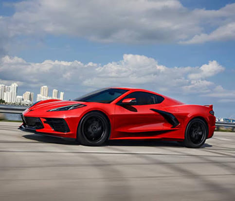 A red Chevrolet Corvette C8 sports car driving on a road with a city skyline in the background.