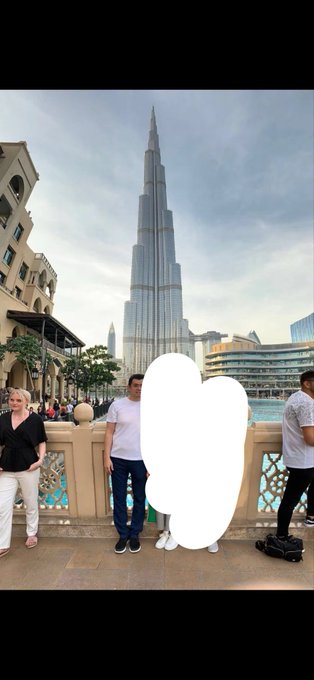 Three people pose for a photo in front of the Burj Khalifa in Dubai, with the central figure obscured by a white cloud. The towering skyscraper dominates the skyline behind them as they stand near the fountain area. The luxurious Dubai setting visually amplifies the post's mocking tone about someone being "the richest guy out."