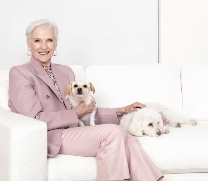 Maye Musk, a brand ambassador for OKX's X Layer, sits smiling on a white couch with her two small dogs, embodying the promotional imagery behind the $CALI meme coin launch tied to tokenized stock rewards on X Layer.